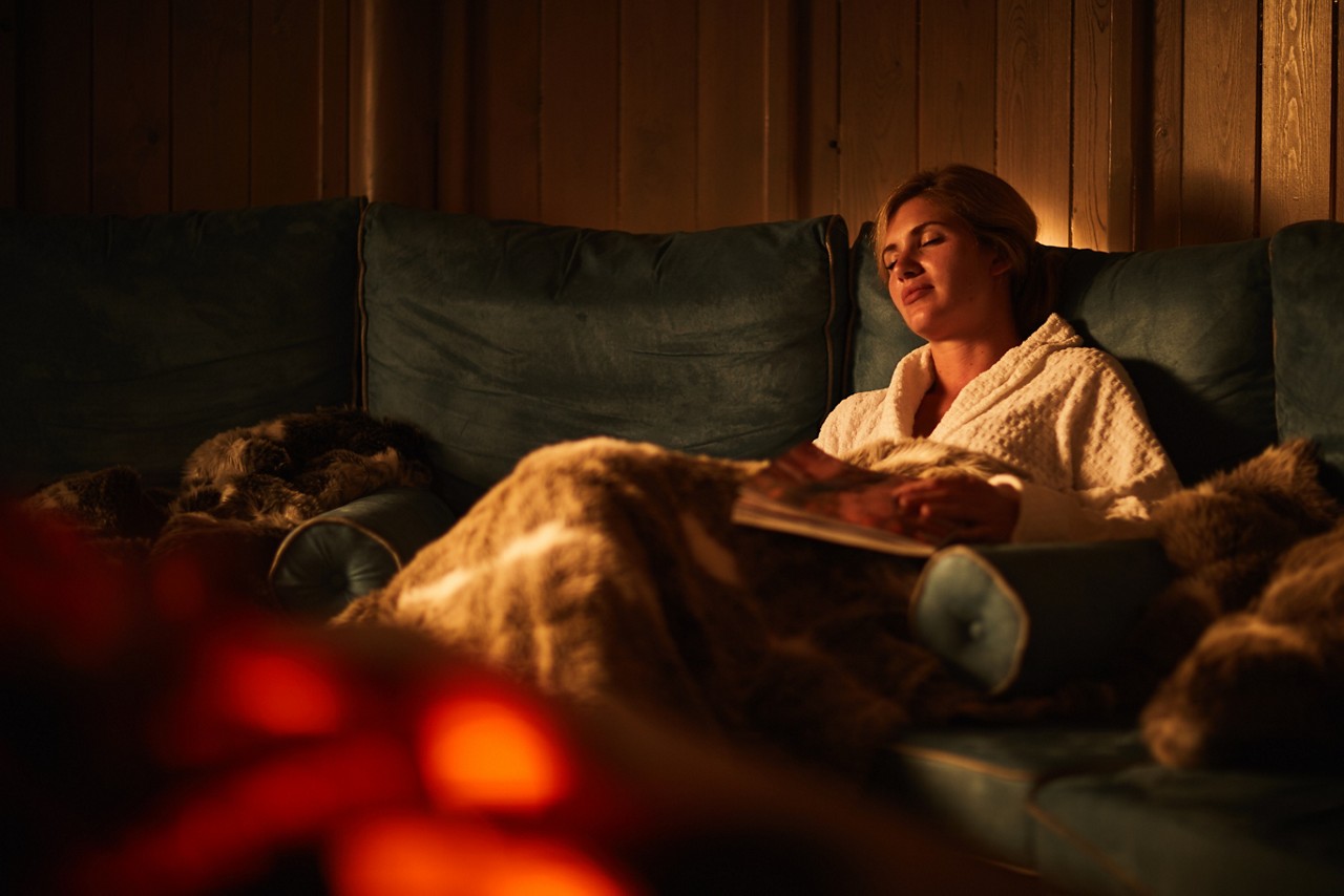 Person reclines on a teal sofa, eyes closed, holding a magazine under a furry blanket; warm firelight flickers in foreground within a wood-paneled, dim, cozy room.
